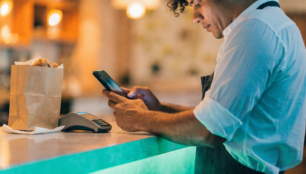 Dono de restaurante no balcão conferindo o celular ao lado de sacola de delivery e maquininha, detalhe turquesa