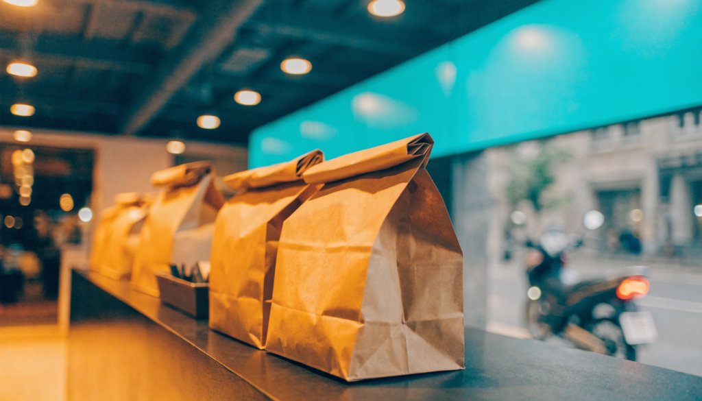 Balcão de restaurante com sacolas de delivery prontas e moto de entrega ao fundo, detalhe turquesa