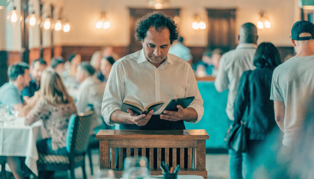 Recepcionista de restaurante no balcão de entrada com livro de reservas e salão cheio ao fundo